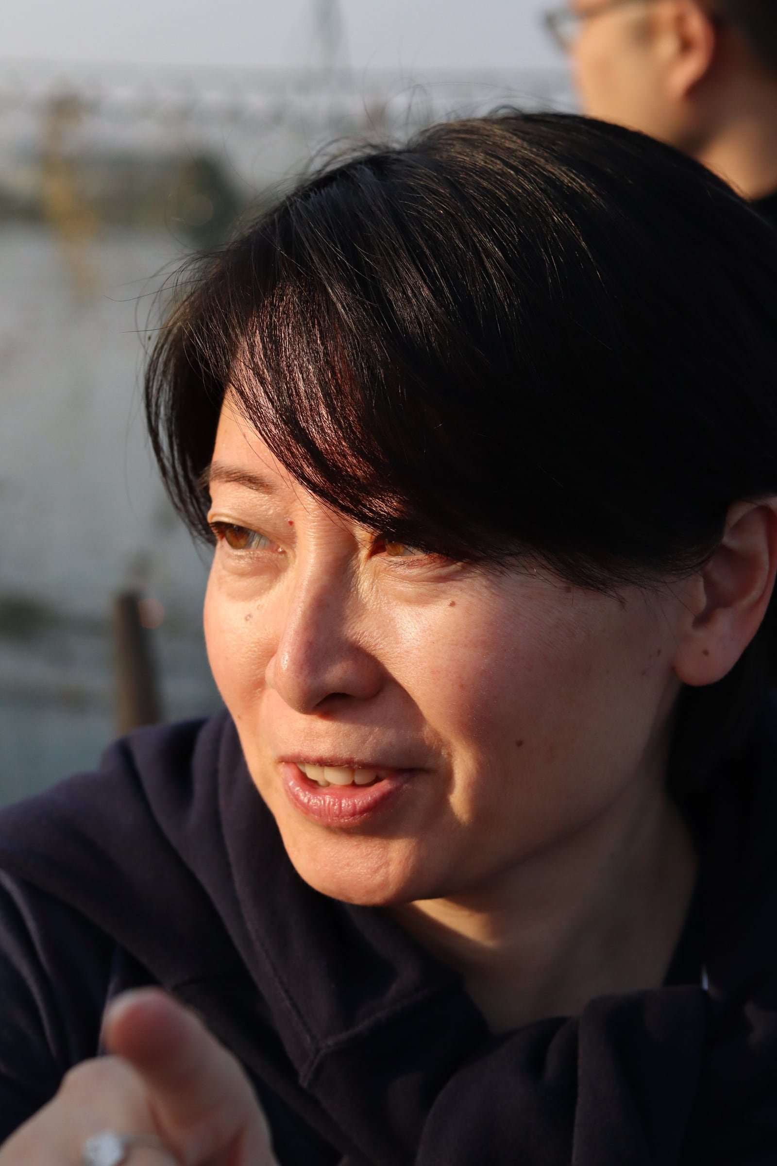 A woman with short dark hair, mid-conversation and pointing, lit by warm afternoon light.