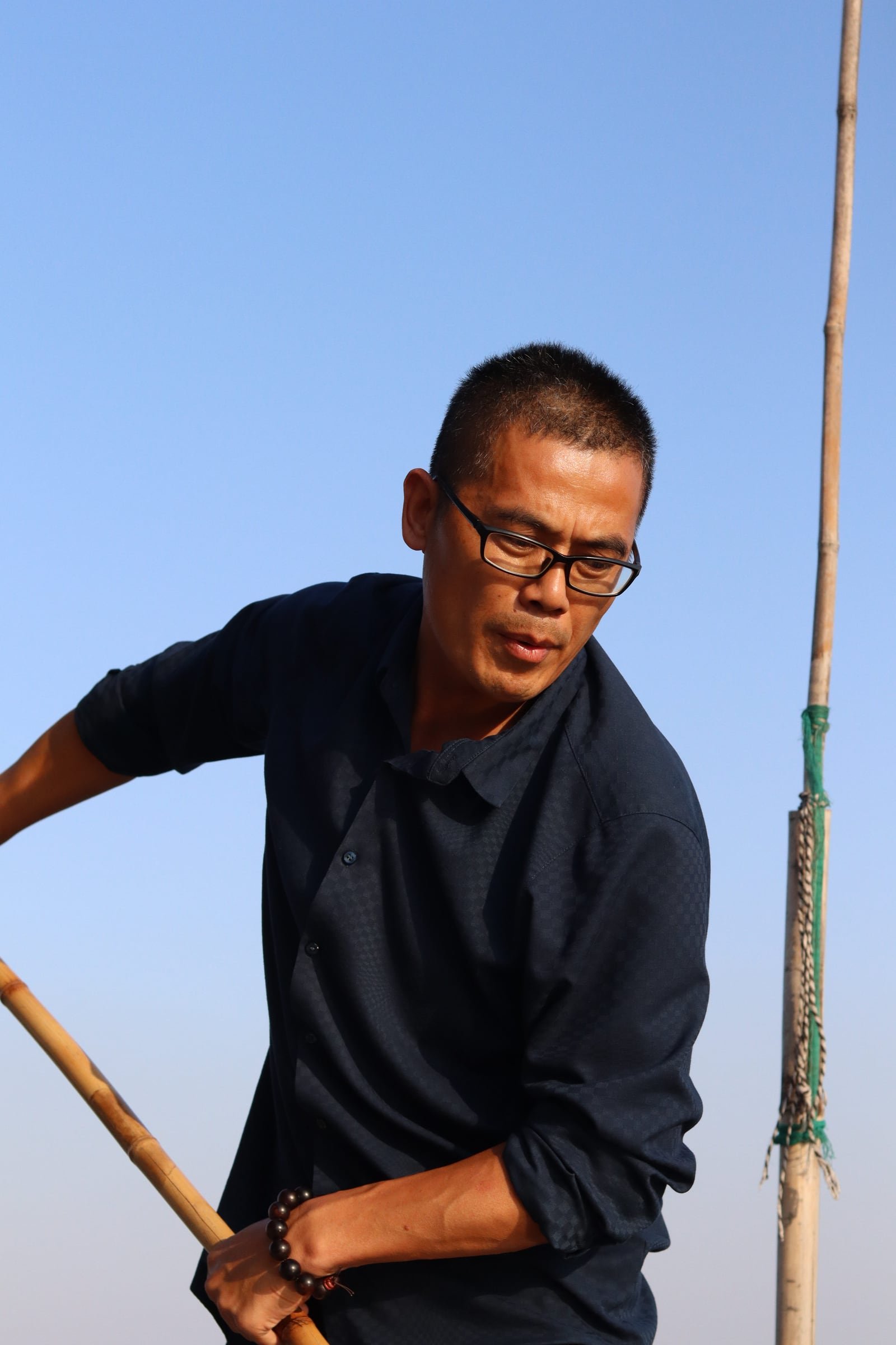 A fisherman in glasses and a navy shirt steering with a bamboo pole against a clear blue sky.