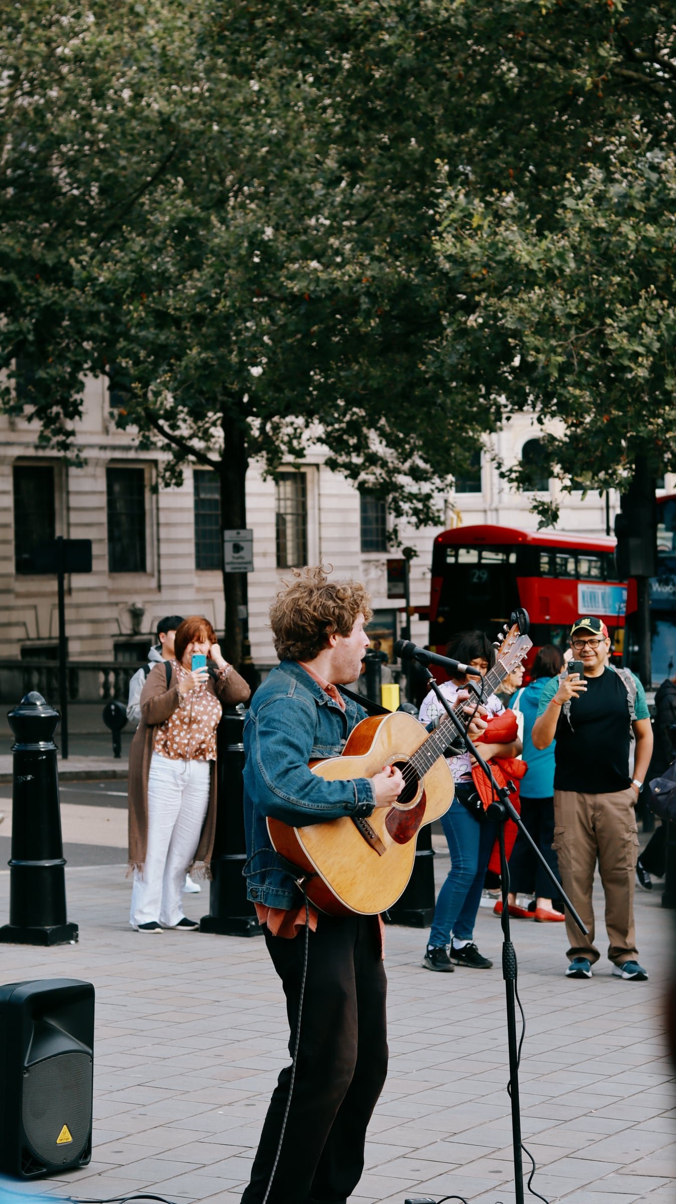 A busker singing with an acoustic guitar in a London square, a red double-decker bus behind him.